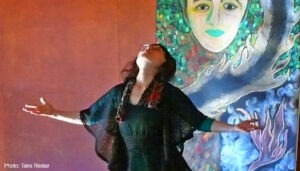 Woman dressed in black dress lifting face to sky with arms open in front of self portrait drwaing of face and blue and purple flames