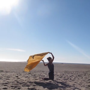 woman's silhouette kneeling in sand at the beach with arms overhead holding a flowing yellow cloth billowing in the wind behind her