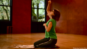 redhaired woman kneeling in wooden dance studio with one arm extended overhead and another cupped by her heart. Look of curiosity on face. Drawing laid out in front of her.