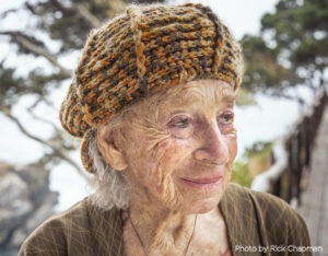 Anna Halprin at age 99, white hair, soft smile in an orange brown beret hat and matching sweater